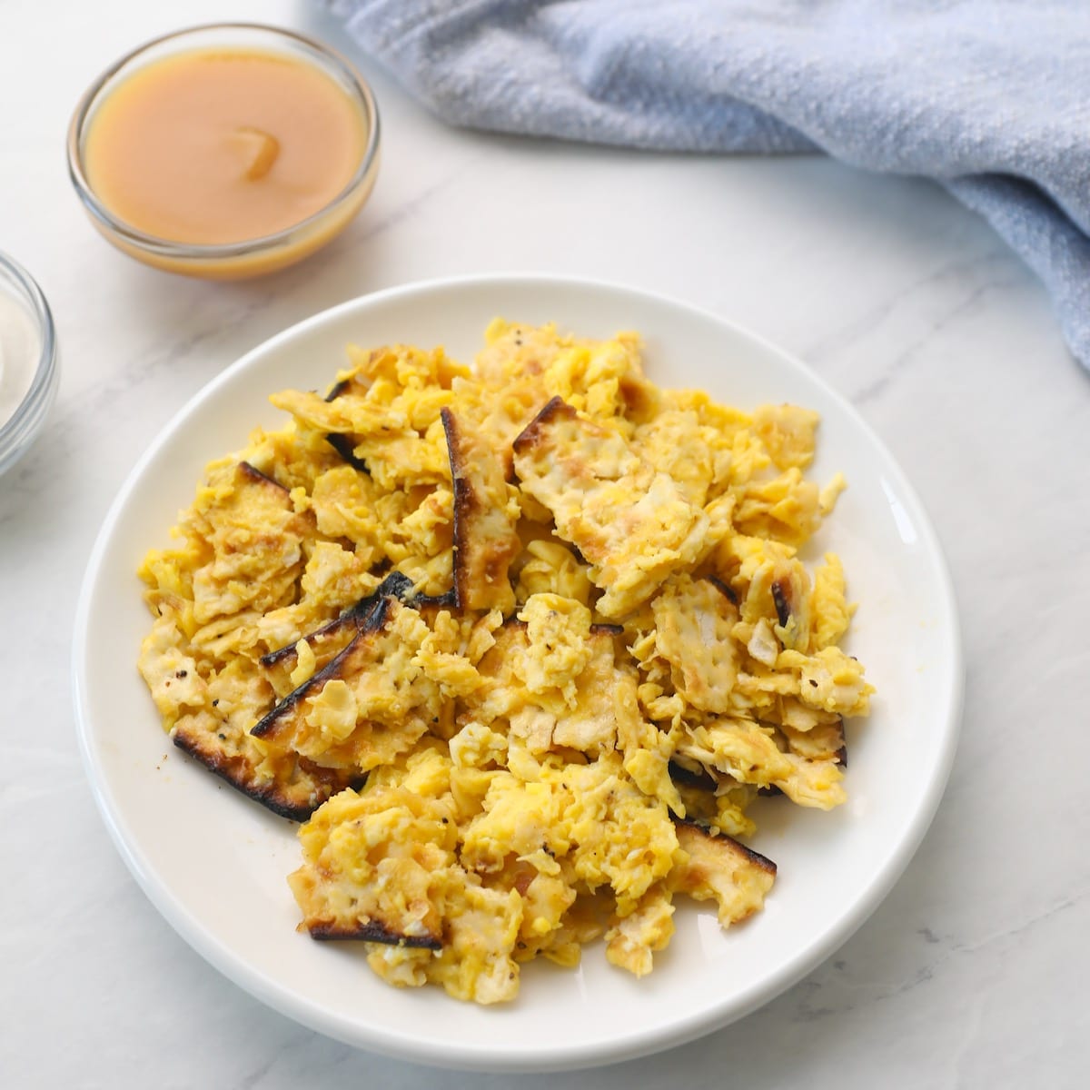 Square featured shot - a white plate with matzo brei eggs and matzo scrambled, small dishes of applesauce and sour cream beside, with a blue towel in background, on a white marble counter.