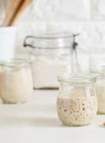 Three small glass jars of sourdough starter in various stages of ferment, with a larger jar of flour and a wooden dish of flour in background. Small wooden spoon and metal measuring cup in foreground, white container of wooden utensils in background, against a white brick wall.