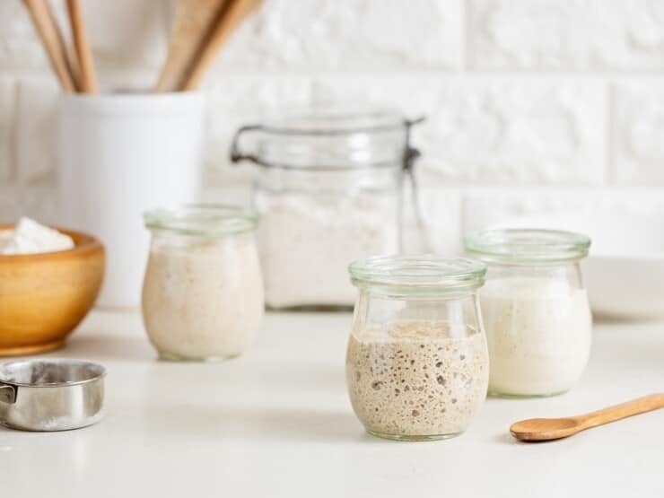 Three small glass jars of sourdough starter in various stages of ferment, with a larger jar of flour and a wooden dish of flour in background. Small wooden spoon and metal measuring cup in foreground, white container of wooden utensils in background, against a white brick wall.