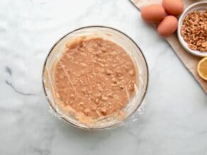 Horizontal overhead shot of a glass mixing bowl containing mixed mandel bread ingredients. A sheet of plastic wrap is laying over the top.