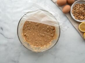 Horizontal overhead shot of a glass mixing bowl containing mixed mandel bread ingredients. A sheet of plastic wrap is laying over the top.