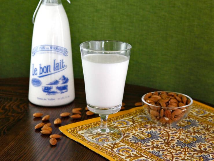 A horizontal image featuring a glass of creamy Almond Milk, with a bowl overflowing with fresh almonds on the left and a bottle of milk on the right.