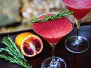 Overhead angled shot of two martini glasses filled with a bright red blood orange cocktail. Both are garnished with a sprig of fresh rosemary. A sliced blood orange and more rosemary sit off to the left.