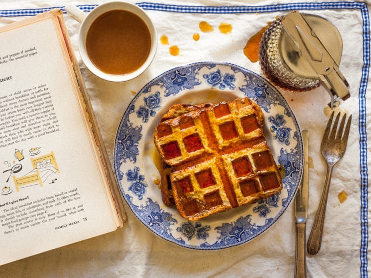 Close-up of two Bread and Butter Waffles soaked on syrup served on a fancy plate with a cup of coffee, syrup glass, and cook book on the side