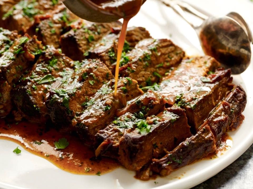 Ladle spooning sauce over sliced brisket topped with fresh parsley on white dish, tongs in background.