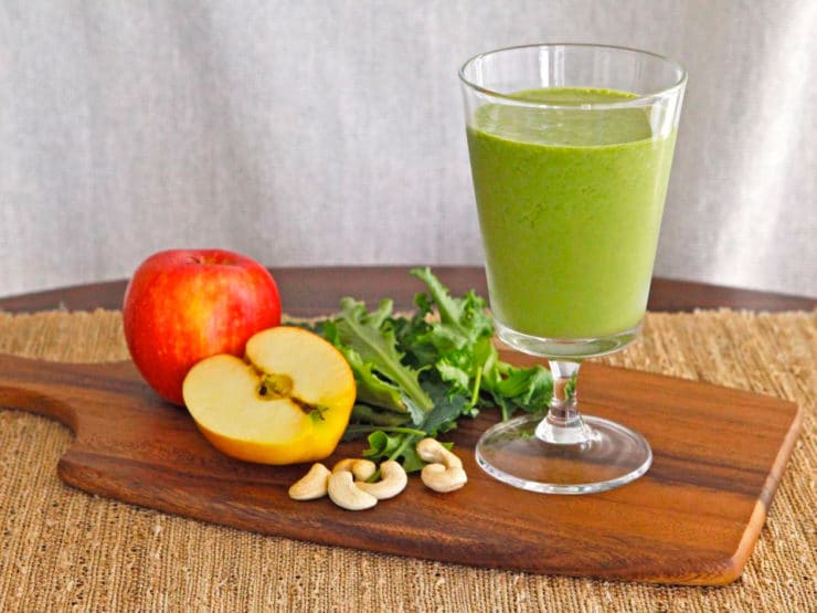 Glass filled with Cashew Apple Green Smoothie surrounded with apples, cashews, and kale