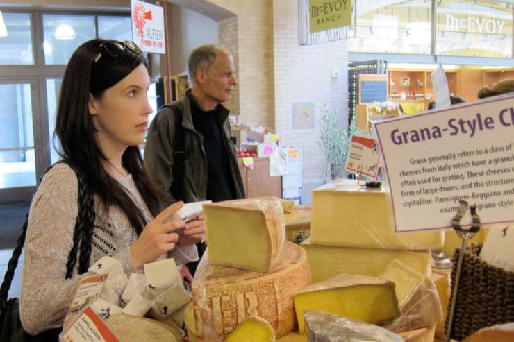 Tori Avey standing at cheese counter at Ferry Plaza Farmer\'s Market, shopper in background.