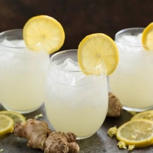 Three glasses of ginger beer with lemon slices on top, ginger and additional lemon slices on sides.