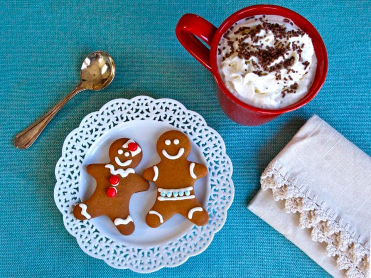 Top view of two gingerbread cookies served on a white fancy plate surrounded with a white table cloth, spoon, and a chocolate drink topped with whipped cream and chocolate sprinkles