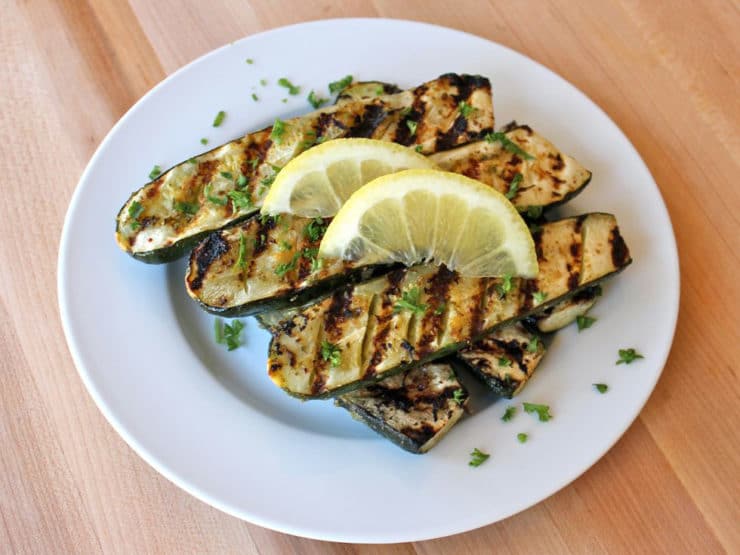 Grilled Lemon Butter Zucchini served on a white plate with 3 slices of lemon and chopped herbs