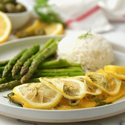 Square image of neatly arranged sliced lemon on a slant, accompanied by a serving of rice in the background and a side of asparagus, all presented on a white round plate