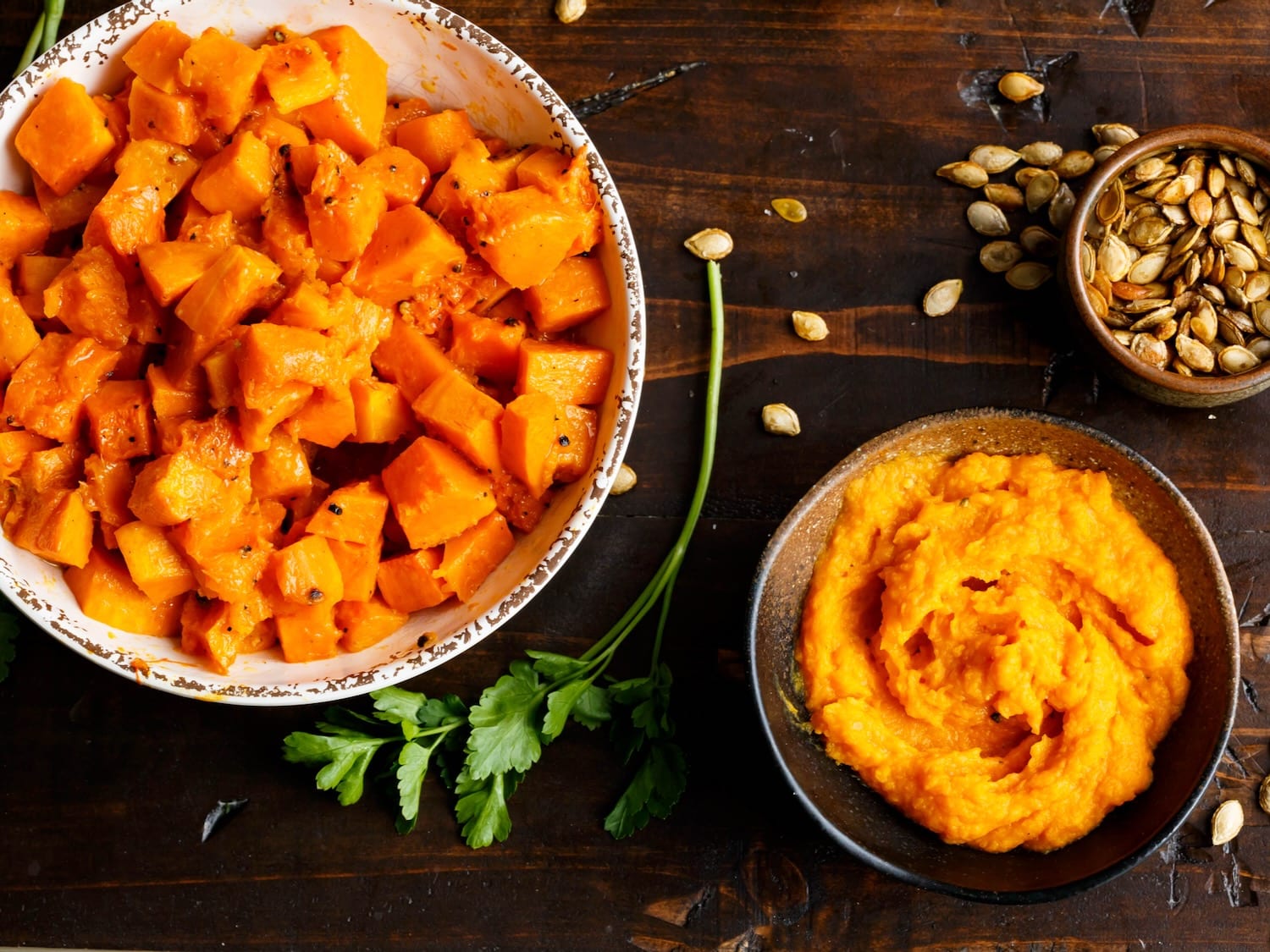 Overhead shot - a large bowl of tender roasted butternut squash cubes, a medium bowl filled with butternut squash puree, and a small bowl filled with toasted butternut squash seeds, on a dark wooden background. Fresh parsley and toasted butternut squash seeds are scattered beside the bowl.