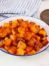 Maple Roasted Butternut Squash in a large serving bowl on a marble countertop. Blue and white linen striped cloth towel and dark wooden spoon beside the bowl.