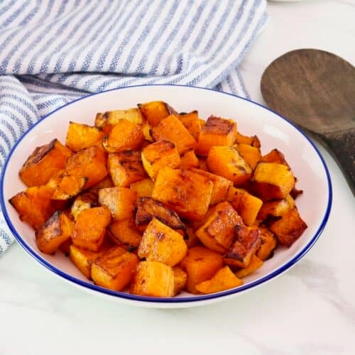 Maple Roasted Butternut Squash in a large serving bowl on a marble countertop. Blue and white linen striped cloth towel and dark wooden spoon beside the bowl.