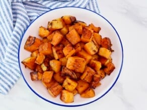 Overhead shot - large bowl of roasted butternut squash, caramelized. Dish sits on a white marble countertop with blue and white linen towel.