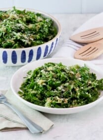 Horizontal shot - dish and serving bowls filled with kale Caesar salad on a white marble countertip. Linen napkins, metal fork, and two wooden serving spoons beside them.