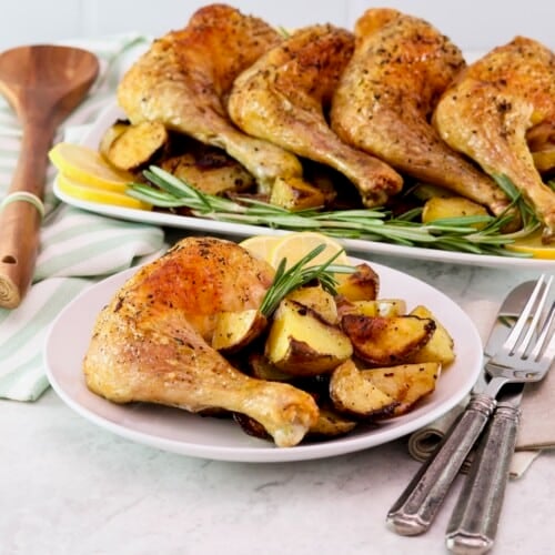 Horizontal shot - in foreground, a small plate with a whole roasted chicken leg, roasted potatoes, and rosemary with, fork, knife, and linen napkin beside it. In background, a platter of roasted chicken legs on a bed of roasted potatoes, lemon, and fresh rosemary. Wooden spoon and kitchen towel beside the platter.