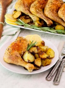 Vertical shot - in foreground, a small plate with a whole roasted chicken leg, roasted potatoes, and rosemary with a fork and knife beside it. In background, a platter of roasted chicken legs on a bed of roasted potatoes, lemon, and fresh rosemary. Wooden spoon and linen towel beside the platter.