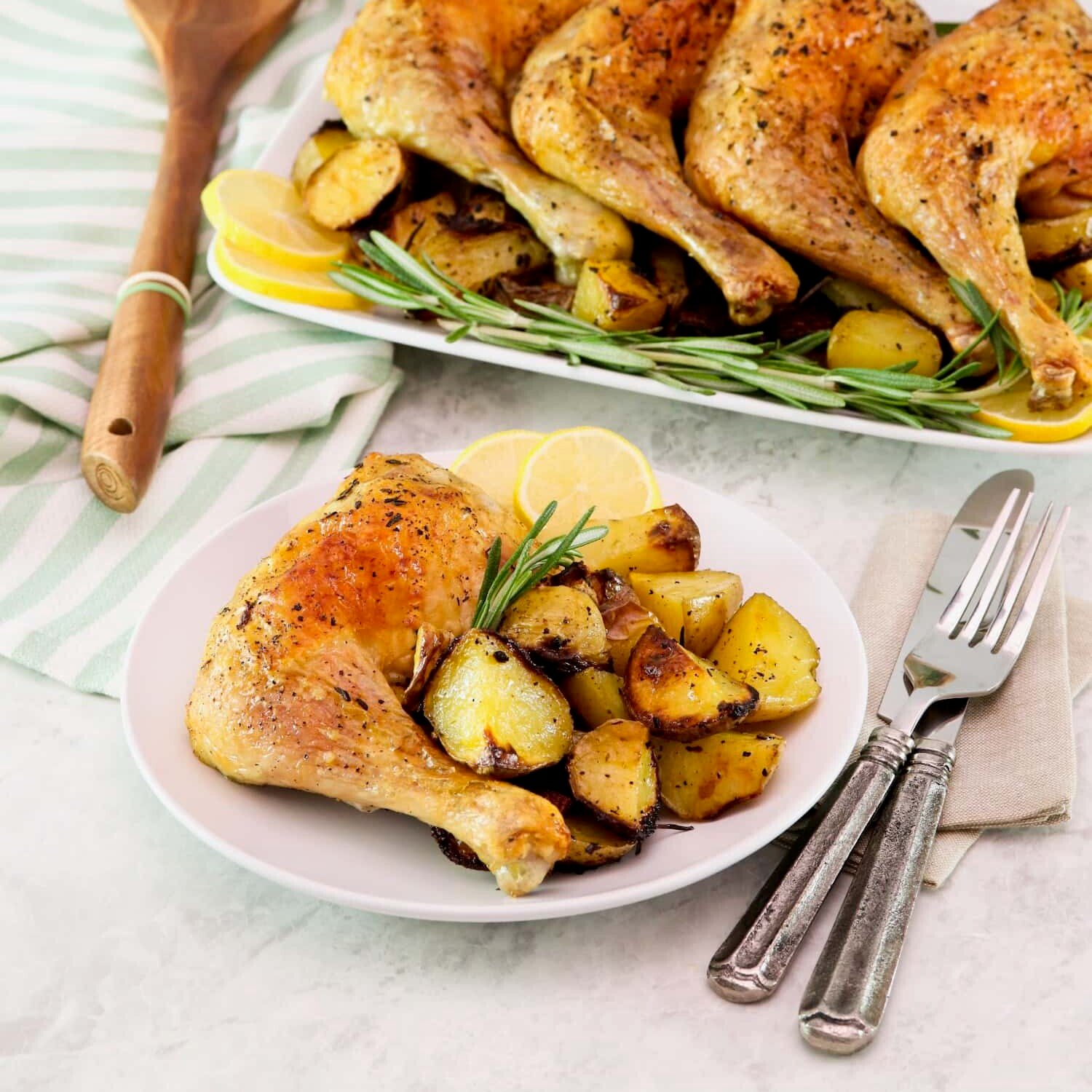 Vertical shot - in foreground, a small plate with a whole roasted chicken leg, roasted potatoes, and rosemary with a fork and knife beside it. In background, a platter of roasted chicken legs on a bed of roasted potatoes, lemon, and fresh rosemary. Wooden spoon and linen towel beside the platter.