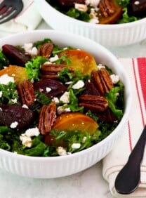 Horizontal shot - shallow bowl filled with roasted beet salad with kale and goat cheese. Fork and striped linen napkin beside the bowl. Partial view of another salad bowl filled with salad, fork, and napkin in background. All resting on a white marble countertop.