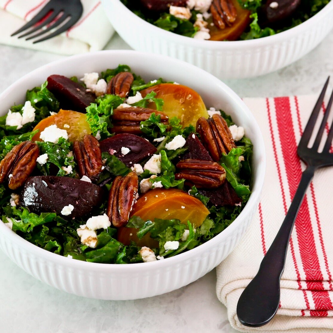 Horizontal shot - shallow bowl filled with roasted beet salad with kale and goat cheese. Fork and striped linen napkin beside the bowl. Partial view of another salad bowl filled with salad, fork, and napkin in background. All resting on a white marble countertop.