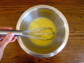 Hand using a whisk to scramble eggs in a large mixing bowl, which rests on a wooden table.