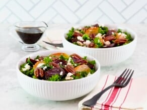 Two shallow bowls filled with roasted beet salad with kale and goat cheese. Fork and linen striped napkin in foreground, small glass pitcher of maple balsamic dressing in background.