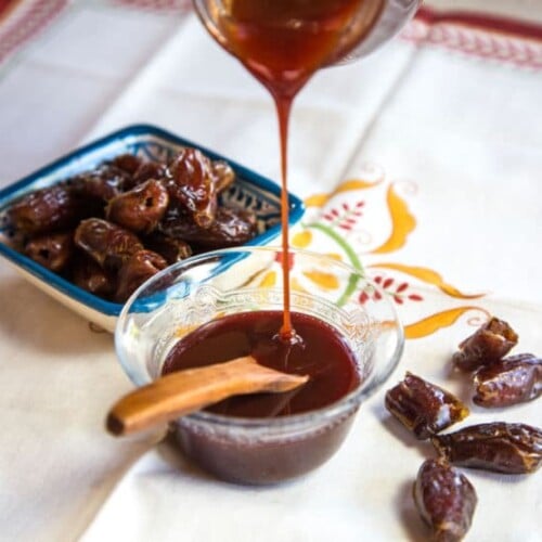 Date Honey Syrup being poured into a glass bowl with wooden spoon, dish of dates in background, scattered dates to the side, on a decorative cloth napkin.