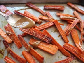 Spoon drizzling roasted carrots with tahini glaze on baking sheet.