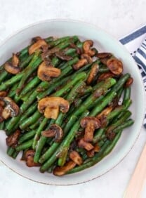 Overhead horizontal shot - large shallow bowl filled with green beans and mushrooms tossed in a rich brown plum sauce. Dish rests on a marble countertop, wooden spoon and kitchen towel beside it.