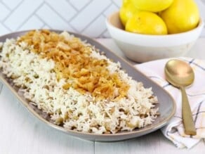 Oblong platter of mujadara topped with caramelized onions. Spoon and cloth napkin beside it. Dish of three lemons in background, against a tile wall.