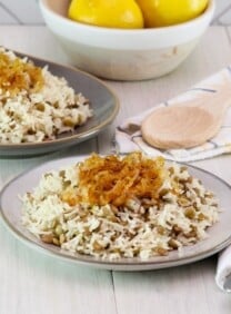 Horizontal shot. In foreground, small dish of mujadara lentils and rice topped with caramelized onions, with fork and cloth napkin beside it. In background, oblong platter of mujadara topped with caramelized onions, spoon and cloth napkin beside it; dish of three lemons in background, against a tile wall.