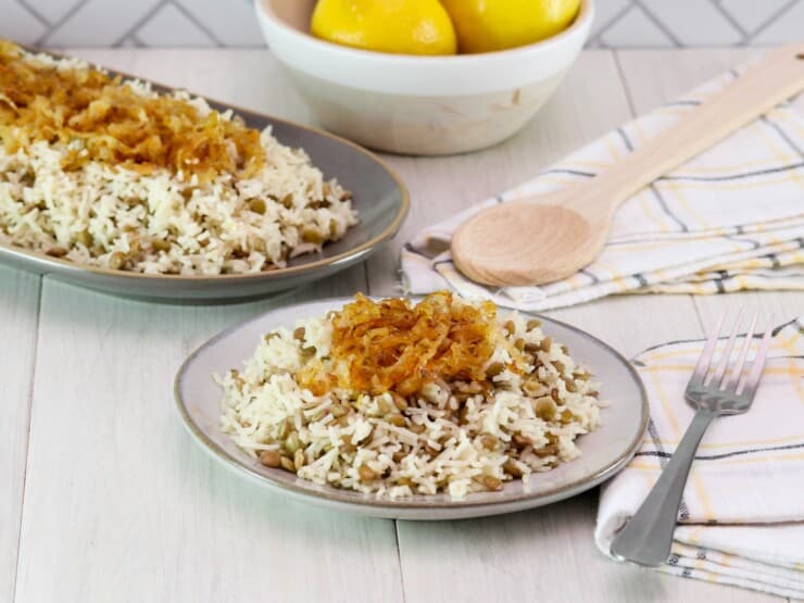 Horizontal shot. In foreground, small dish of mujadara lentils and rice topped with caramelized onions, with fork and cloth napkin beside it. In background, oblong platter of mujadara topped with caramelized onions, spoon and cloth napkin beside it; dish of three lemons in background, against a tile wall.