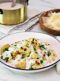 Horizontal shot - large shallow bowl of roasted cauliflower Alfredo with peas, covered in a rich Greek yogurt Alfredo sauce, on a marble countertop. Fork and napkin with green stripes beside the bowl. Small wooden bowl of shaved parmesan in upper right corner of the picture, and a small gold metal pan of Alfredo sauce with freshly cracked black pepper in upper left hand corner. Gold pan rests on a white linen towel.