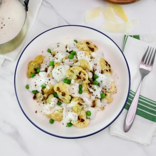 Overhead shot - large shallow bowl of roasted cauliflower alfredo with peas, covered in a thick Greek yogurt Alfredo sauce, on a marble countertop. Fork and napkin with green stripes beside the bowl. Shaved parmesan and a small metal pan of Alfredo sauce with freshly cracked black pepper in upper left hand corner.