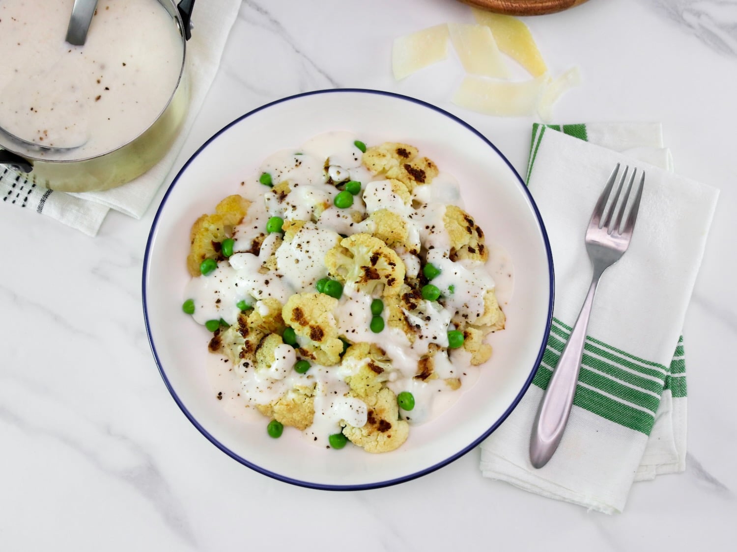 Overhead shot - large shallow bowl of roasted cauliflower alfredo with peas, covered in a thick Greek yogurt Alfredo sauce, on a marble countertop. Fork and napkin with green stripes beside the bowl. Shaved parmesan and a small metal pan of Alfredo sauce with freshly cracked black pepper in upper left hand corner.