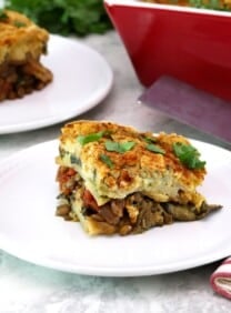 A square slice of vegan moussaka on a small white plate in foreground with fork and linen napkin. A red, rectangular baking dish and a stainless steel serving shovel on the background with another serving of a sliced vegan moussaka.