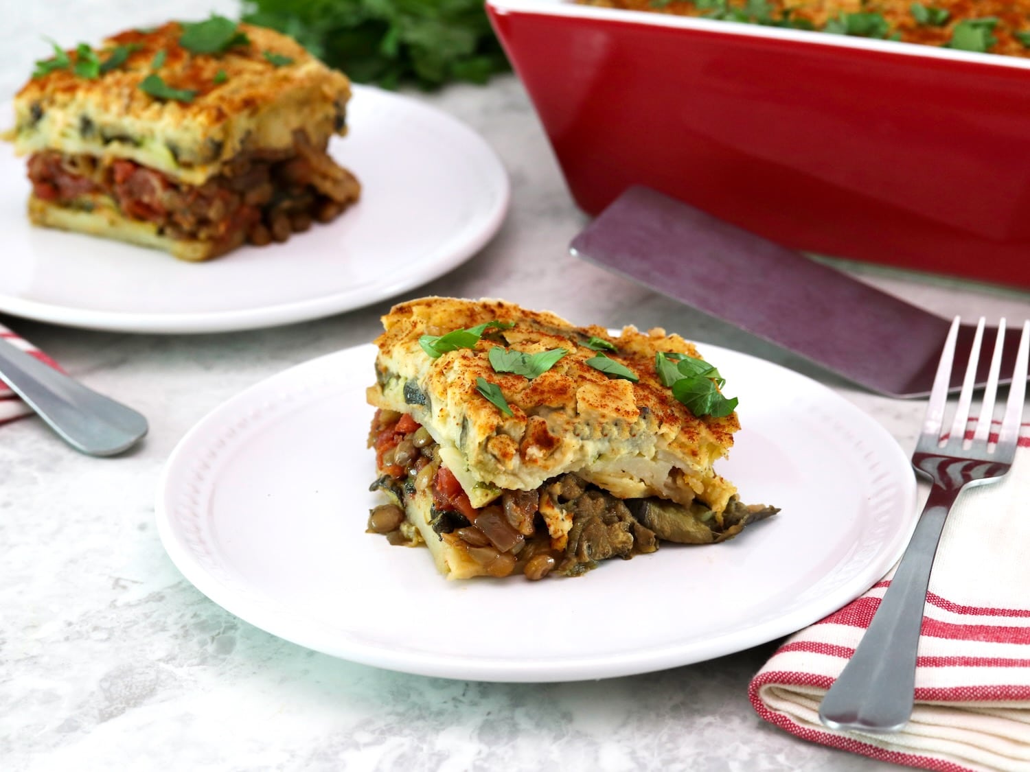 A square slice of vegan moussaka on a small white plate in foreground with fork and linen napkin. A red, rectangular baking dish and a stainless steel serving shovel on the background with another serving of a sliced vegan moussaka.