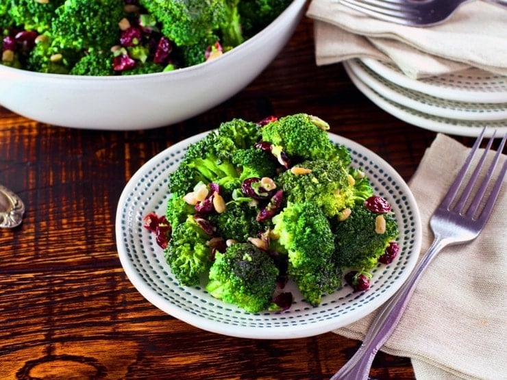 A plate of broccoli salad next to a fork and napkin on a wooden table. In the background is a large bowl of additional broccoli salad.