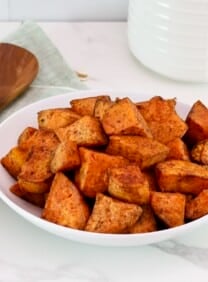 A serving of curry roasted sweet potatoes on a white bowl in foreground with a wooden spoon and linen napkin on the left side. White canister in the background.