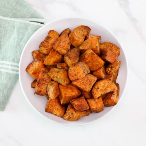 A serving of curry roasted sweet potatoes on a white bowl in foreground with a linen napkin on the left side.