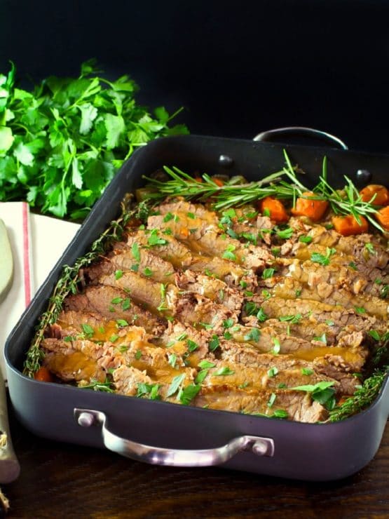 Roasting pan with cooked sliced brisket and carrots topped with fresh herbs, fresh parsley and cloth napkin on the side.