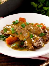 White plate with tender slices of savory herb braised brisket and carrots, topped with fresh herbs. Napkin, fork and fresh herbs in background with roasting pan of meat.