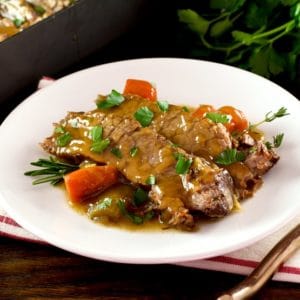 White plate with tender slices of savory herb braised brisket and carrots, topped with fresh herbs. Napkin, fork and fresh herbs in background with roasting pan of meat.