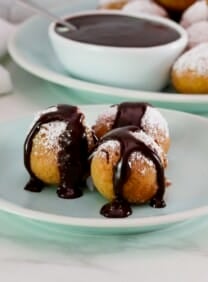 Small dessert plate of three fried buñuelos or bimuelos, topped with powdered sugar and drizzled with chocolate sauce. Platter of powdered sugar buñuelos and a dish of chocolate sauce, cloth napkin in background.