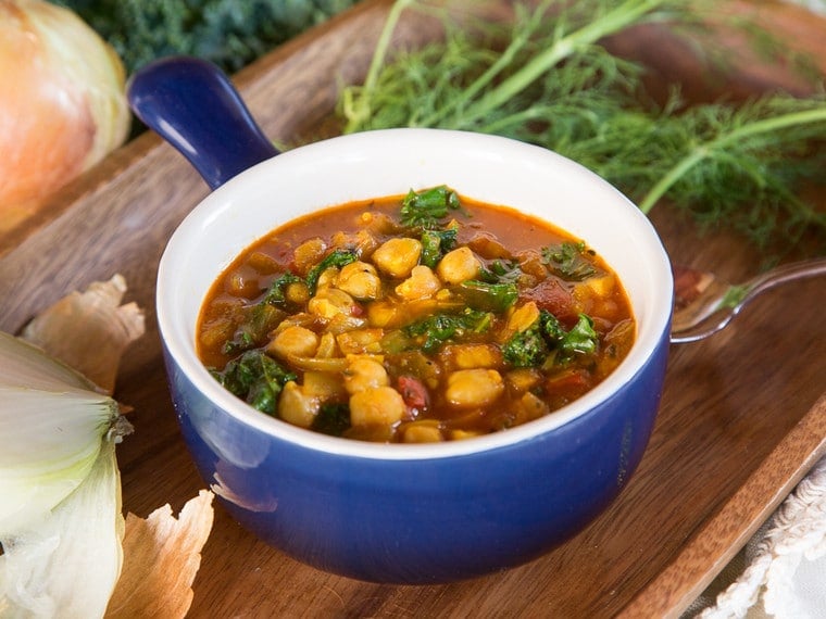 A bowl of Chickpea Kale and Fire Roasted Tomato Soup with vegetables and herbs on a cutting board