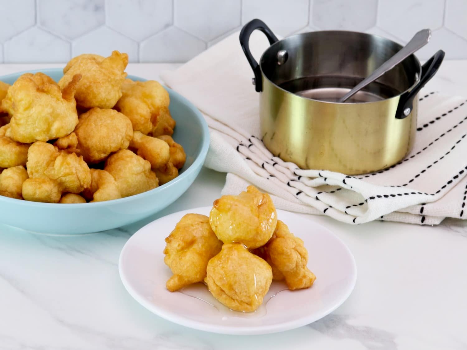 Horizontal shot - Hanukkah buñuelos dessert - pile of four on a small white plate drenched in clear syrup. Large plate of buñuelos in background, as well as a gold pot of clear sauce, on a white and blue linen towel.