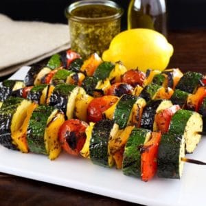 Four lemon pesto vegetable skewers on white plate on wooden table, lemon, pesto and oil in background with napkin.