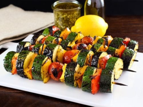 Four lemon pesto vegetable skewers on white plate on wooden table, lemon, pesto and oil in background with napkin.