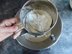 Female hand sifting flour over a mixing bowl on concrete surface.